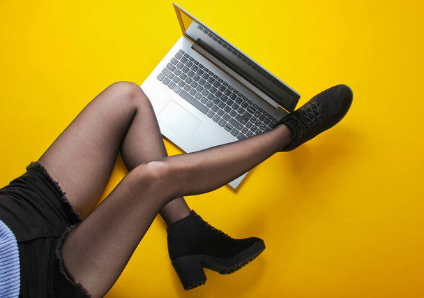 Modieuze vrouw in shorts, panty 's en laarzen gebruiken laptop terwijl ze op een gele achtergrond zit. Freelance, blogger, online werknemer concept. Bovenaanzicht - Foto, afbeelding