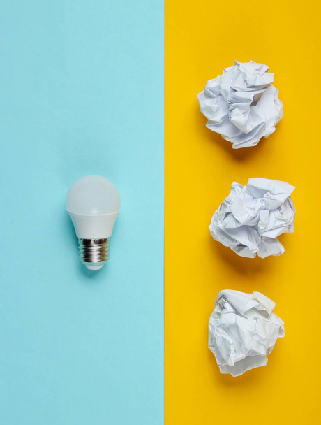 Energy saving LED light bulb and crumpled paper balls on yellow blue background. Minimalistic business concept, idea. Top view - 写真・画像
