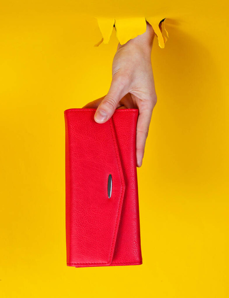 Female hand holding a red wallet through torn yellow paper. Minimalistic creative fashion concept - Foto, afbeelding