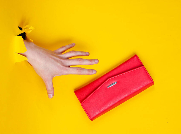 A női kéz átveszi a piros erszényt a szakadt sárga papíron. Minimalista kreatív divat koncepció - Fotó, kép