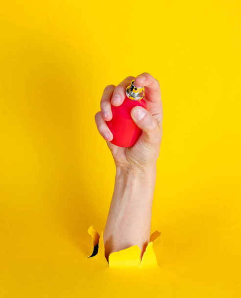 Botella de mano femenina de perfume a través de papel amarillo roto. Concepto de moda minimalista
 - Foto, imagen