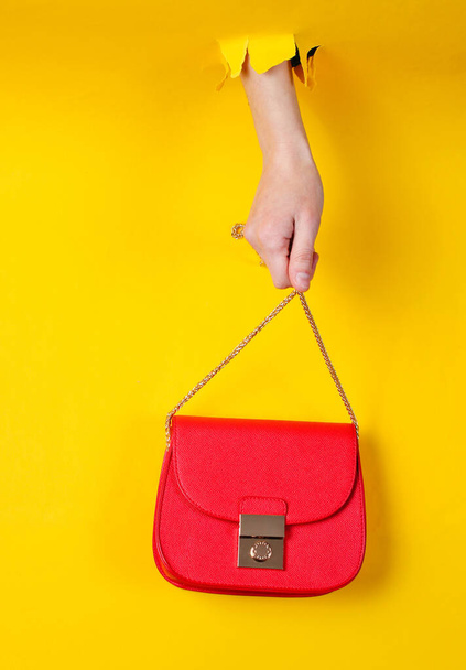 Female hand is holding red leather bag over chain through torn yellow paper. Minimalistic fashion concept - 写真・画像