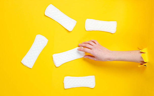 Female hand holds daily sanitary pad through torn yellow paper background. Minimalistic fashion concept - Photo, Image