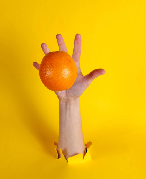 Θηλυκό χέρι ρίχνει πορτοκαλί μέσα από το σκισμένο κίτρινο φόντο χαρτί. Μινιμαλιστική έννοια φρούτων - Φωτογραφία, εικόνα