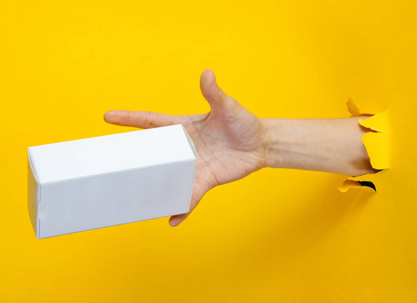 Female hand catches white box through torn yellow paper. Minimalistic creative concept - 写真・画像