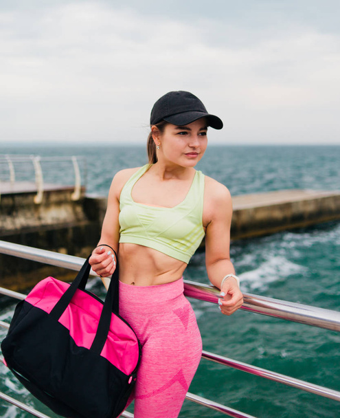 Spor kıyafetli genç bir kadın, elinde eğitim çantası ve sahilde poz vermek için bir çanta tutuyor. - Fotoğraf, Görsel