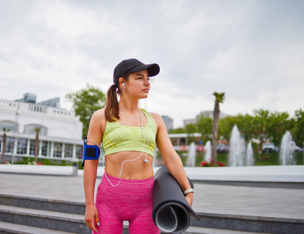 Athlétique jeune femme en tenue de sport tient un tapis de yoga dans sa main et écoute de la musique avec casque à l'extérieur
 - Photo, image