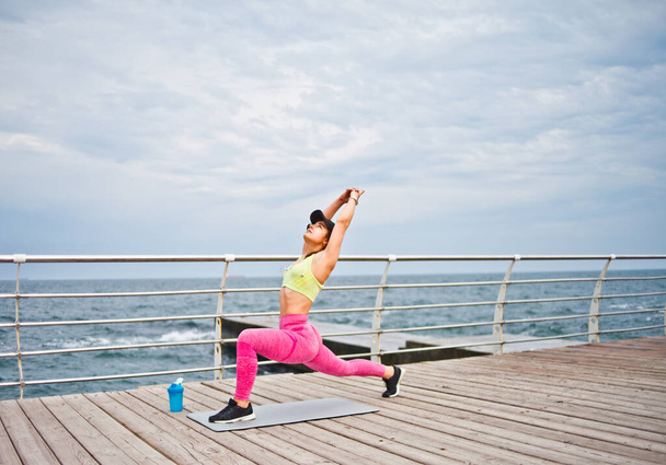 Mladá fit žena ve sportovním oblečení a čepice dělá jóga cvičení na pláži v dopoledních hodinách - Fotografie, Obrázek