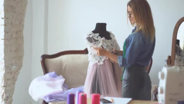 Hermosa costurera rubia en el estudio de costura trabaja con tela cerca de maniquí. El proceso de creación de modelos de ropa de mujer
 - Imágenes, Vídeo