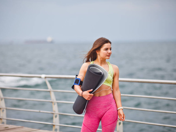 Spor kıyafetli genç bir kadın elinde yoga minderiyle sahilde poz veriyor. - Fotoğraf, Görsel