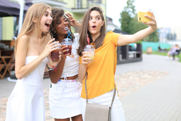 Τρία μοντέρνα cool hipster κορίτσια, οι φίλοι πίνουν κοκτέιλ και τη λήψη selfie στο φόντο της πόλης. - Φωτογραφία, εικόνα
