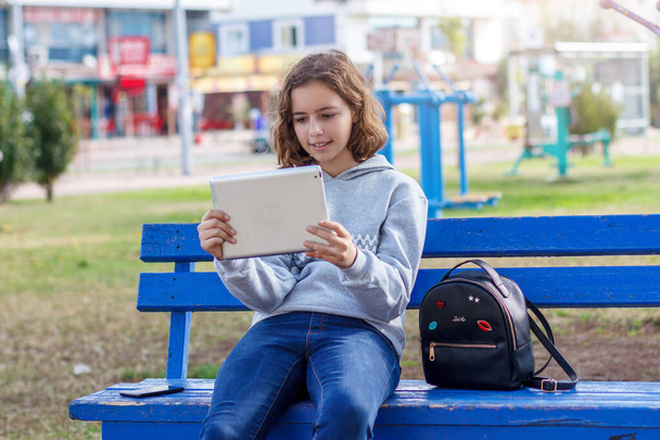 Hermosa chica rizada adolescente viendo un video en una tableta en un parque en un banco
 - Foto, Imagen