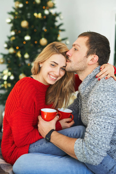 Romantic couple in love embracing, kissing and holding cups with coffee at home with christmas tree on background. - Photo, Image