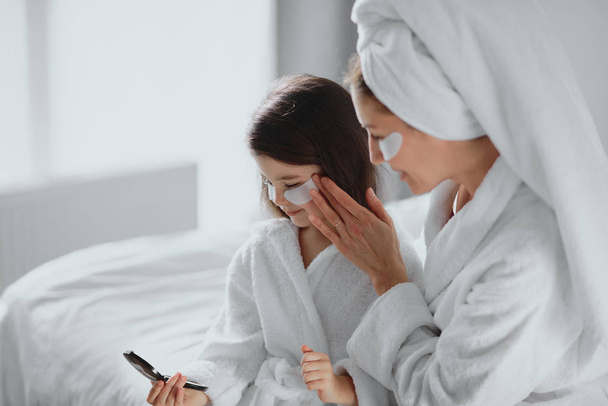 cute pleasant middle-aged mother and her little daughter in bathrobe and mask - Foto, Bild