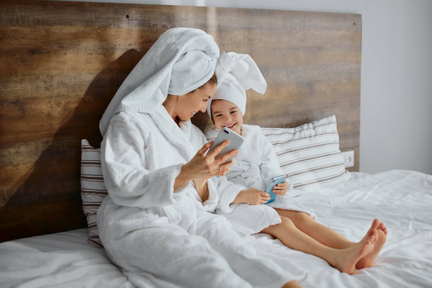attractive woman with daughter lie on bed with mobile phone - Fotoğraf, Görsel