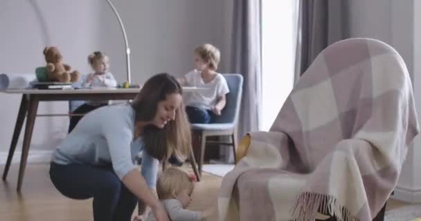 Brunette mother coming and lifting up cute cheerful Caucasian baby girl. Happy young woman taking care of her smallest daughter at the background of other children. Cinema 4k ProRes HQ. - Imágenes, Vídeo