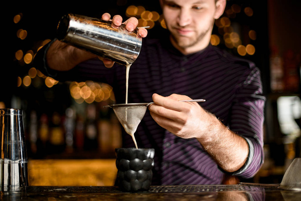 Male bartender pouring a alcoholic drink from the steel shaker to the black cocktail glass through the sieve - Zdjęcie, obraz