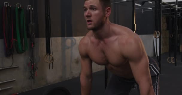 Front view close up of a shirtless muscular Caucasian man cross training at a gym, lifting weights on a barbell, slow motion - Materiał filmowy, wideo