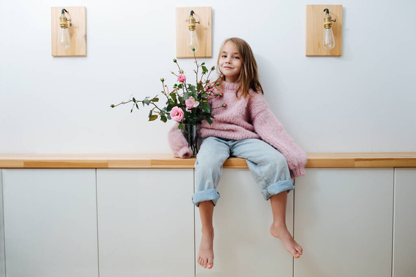 Little barefoot girl in oversized sweater sitting on a shelf hugging flower jar - Fotoğraf, Görsel