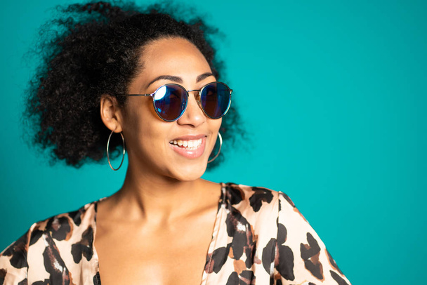 Pretty african woman with afro hair in leopard top and sunglasses smiling to camera over blue wall background. Cute mixed race girls portrait. - Φωτογραφία, εικόνα