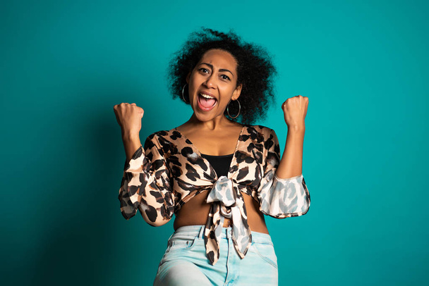 Surprised excited happy african woman on blue background. Girl with curly hair and positive trendy appearance shows yes gesture of victory,she achieved result, goals. - Fotografie, Obrázek