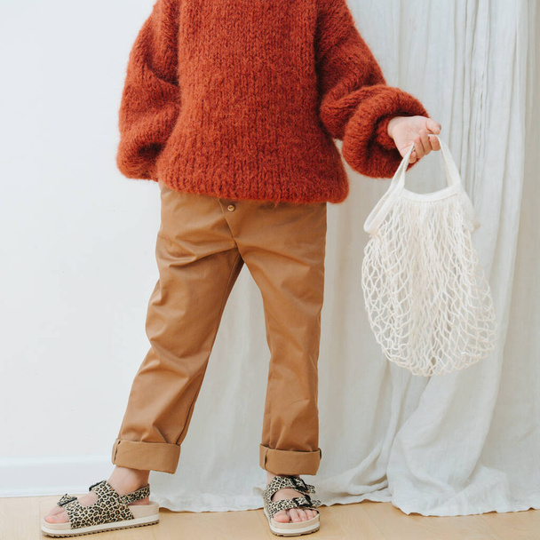 Child in dark orange knitted sweater, brown jeans, sandals with net bag - Photo, Image