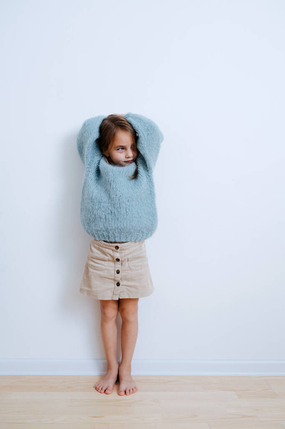 Little barefoot girl in a blue sweater leaning against the wall at home - Valokuva, kuva