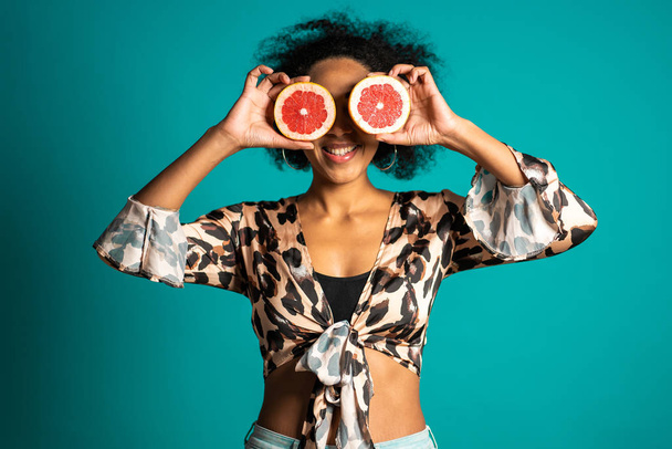 Pretty african american woman in leopard print top with two half of juicy grapefruit on blue studio background. Healthy eating, dieting, antioxidants concept. - Foto, Imagem