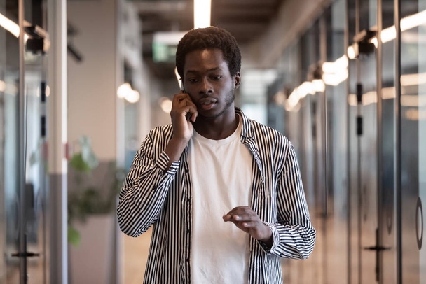 Skoncentrowany afrykański przedsiębiorca spacerujący po korytarzu biurowym rozmawiający przez telefon - Zdjęcie, obraz