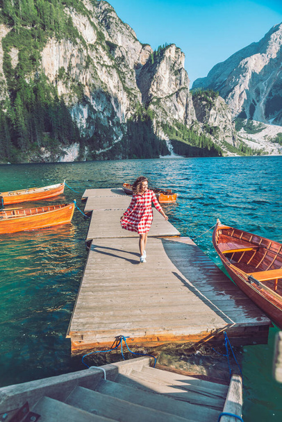 woman walking by mountain lake pier with wooden boats in red dress - Photo, Image
