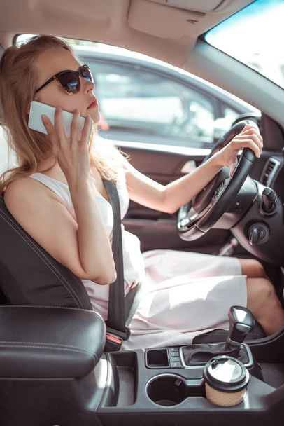 woman driving car in dress, stands in parking lot, looks in rearview mirror, makes phone calls and listens voice messages, reverses, city parking lot of shopping center in summer. - Foto, imagen