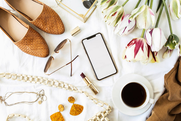 Fashion / beauty blogger home office desk. Blank screen smart phone, tulip flowers bouquet, clothes and accessories on white linen. Flat lay, top view lifestyle composition. - Foto, Bild