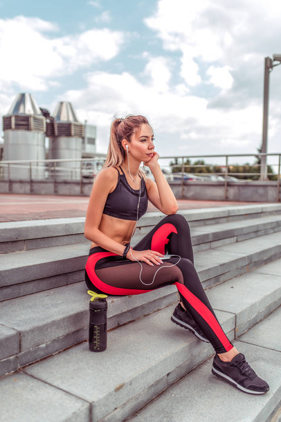 Girl athlete sits on stairs, rest after workout, summer day city. In hand, smartphone listens music headphones. Enjoys listens to podcast lecture. Active lifestyle fitness, fashionable stylish woman. - Photo, image