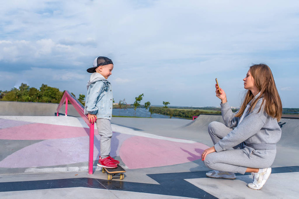 Woman mom takes pictures on phone, support care training, little boy 3-5 years old son stands on skateboard, trains in summer to ride city, autumn on sports ground, happy smiling casual clothes. - Photo, image