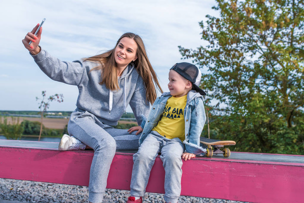 Woman mom takes pictures selfie phone little boy 3-5 years old son, sits resting, summer city autumn sports ground happy smiling casual clothes. Skateboard video call smartphone application Internet. - Photo, image