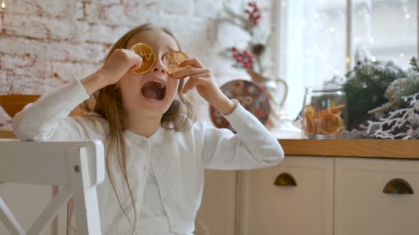 lächelndes kleines blondes Mädchen grimmig und bedeckt ihre Augen mit getrockneten Orangenscheiben zu Hause mit Loft-Stil und Weihnachtsdekoration auf dem Hintergrund, fröhliches Weihnachtskonzept - Filmmaterial, Video