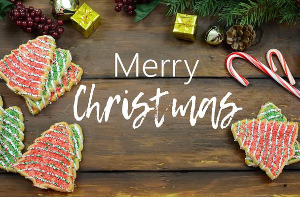 Χριστουγεννιάτικα μπισκότα ζάχαρης σε σχήμα δέντρου, ζαχαρωτά κλαδιά πεύκου με κώνους, μίνι δώρα και κουδουνάκια σε φαρδιά σανίδα ρουστίκ ξύλινο φόντο. Προστέθηκε κείμενο. - Φωτογραφία, εικόνα