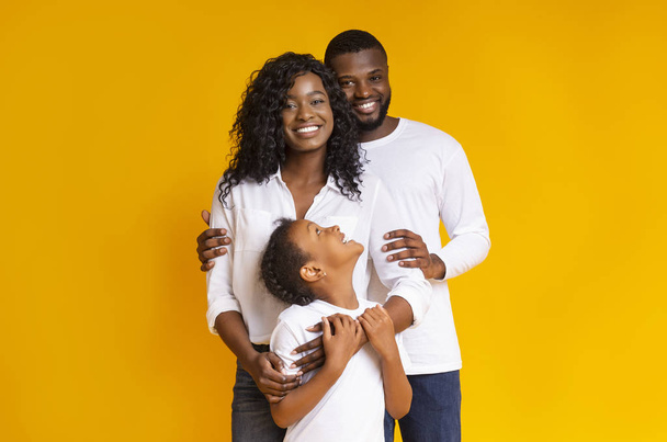 Liefdevolle zwarte familie met dochter omarmen en glimlachen op de camera - Foto, afbeelding