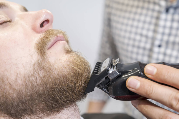 a hairdresser shapes his clients beard with an electric trimmer. The client sits with his eyes closed, selective focus - Photo, Image