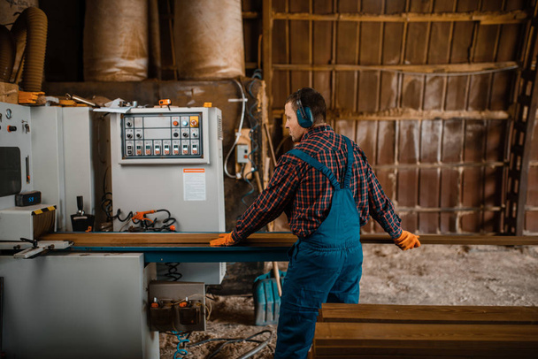 Dřevorubec v uniformě a sluchátka pracuje na dřevozpracujícím stroji, dřevařském průmyslu, tesařství. Továrna na dřevo - Fotografie, Obrázek