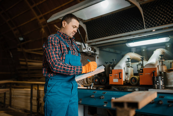 Carpenter in uniform holds notebook, woodworking machine on background, lumber industry, carpentry. Wood processing on factory, forest sawing in lumberyard - 写真・画像