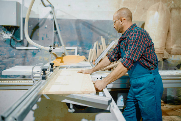 Timmerman snijdt houten plaat op cirkelzaagmachine, houtbewerking, houtindustrie, timmerwerk. Houtbewerking op meubelfabriek, productie van producten van natuurlijke materialen - Foto, afbeelding