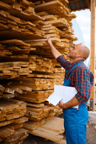 Carpenter in uniform checks boards on sawmill, lumber industry, carpentry. Wood processing on factory, forest sawing in lumberyard, warehouse outdoor - Φωτογραφία, εικόνα