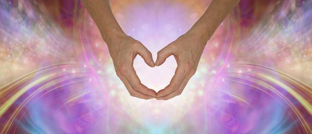 Zend je pure onvoorwaardelijke liefde - handen die een zachte hartvorm maken tegen een etherisch glinsterende veelkleurige energieveld achtergrond met kopieerruimte - Foto, afbeelding