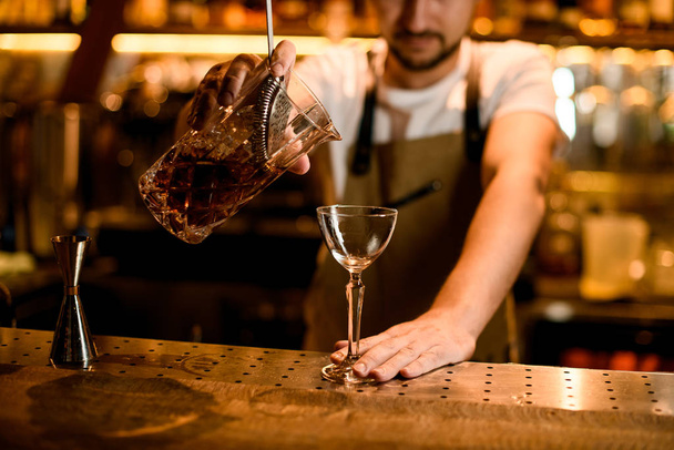 Male bartender pouring a brown alcoholic drink from mixing glass through the sieve - Foto, Bild