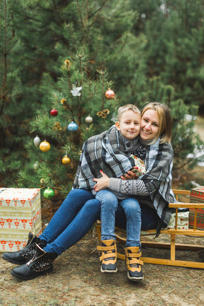 Äiti pienen poikansa kanssa talvipuistossa kelkkailemassa. Äiti ja lapsi istuvat yhdessä puisilla koristelluilla kelkoilla joululahjoilla. Talvi kävellä ulkona, koristeltu joulukuusi
 - Valokuva, kuva