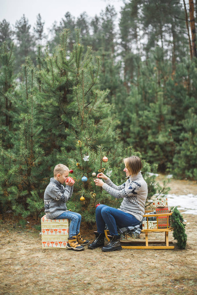 Pretty young mother and the son in gray sweaters decorating a Christmas tree in winter forest outdoors. Boy is drinking hot tea and sitting on wooden sledge with present boxes under the tree - Valokuva, kuva