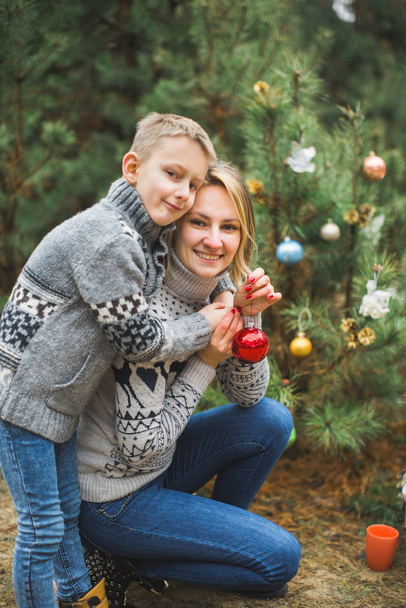 Family, young mother and little son, wearing knitted sweaters, decorating fir tree in the forest or park. Colorful decorations, Christmas presents - Фото, зображення