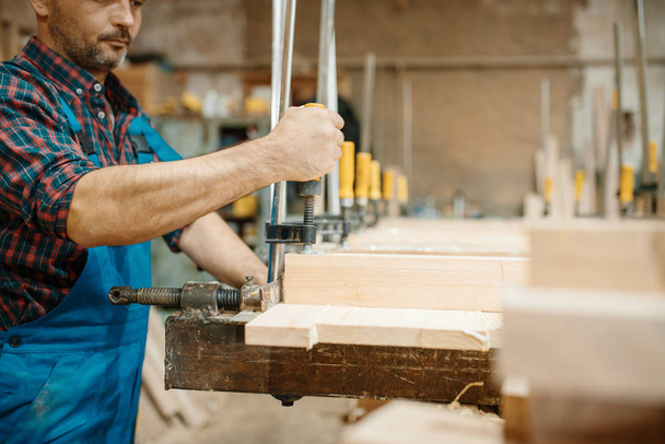 Timmerman in uniform klemt het bord in een bankschroef, houtbewerking, houtindustrie, timmerwerk. Houtbewerking op meubelfabriek, productie van producten van natuurlijke materialen - Foto, afbeelding