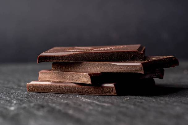 Broken pieces of dark bitter chocolate on black stone surface. - Foto, Bild